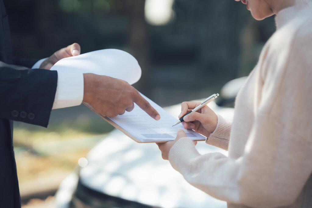 Signing the Documents: Woman signing document with a pen, a man pointing at the document. Close-up view, focus on hands and document. The car accident scene is blurred in the background.